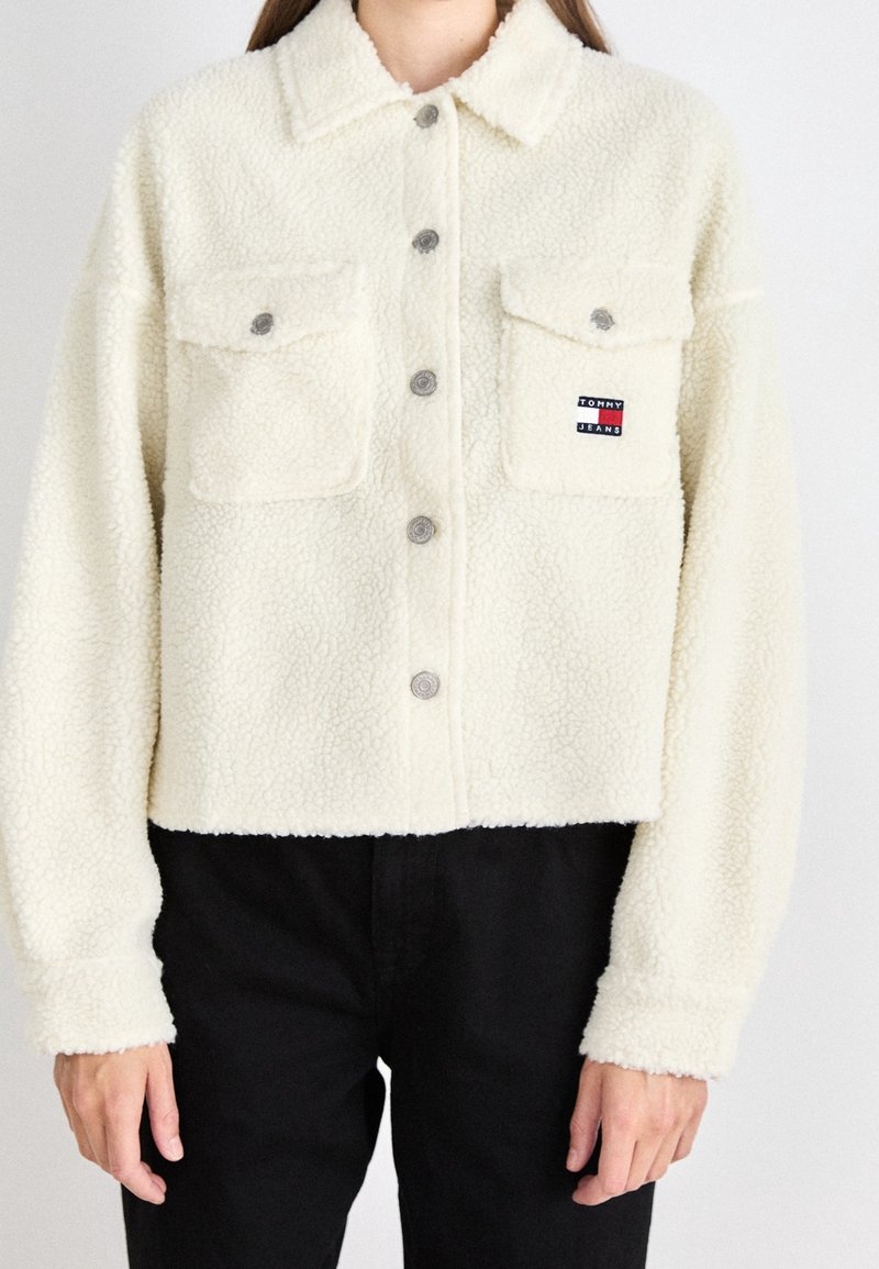Veste courte en polaire blanche avec un front à boutons et deux poches poitrine. Présente des boutons gris et un petit patch logo sur la poche gauche.
