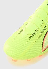 Lyse neon gule fodboldstøvler med tekstureret overflade, sorte accentlinjer og nittede knopper med orange spidser. Har tynde snørebånd på oversiden.