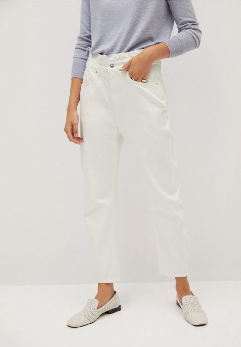 Personne portant un pantalon blanc taille haute à jambes droites, un haut gris clair à manches longues et des chaussures blanches slip-on, main dans la poche.