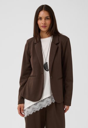 Frau trägt einen braunen Blazer und eine Hose mit einem weißen Spitzen-Top und einer schwarzen Kamm-Anhänger-Halskette vor einem einfarbigen Hintergrund.