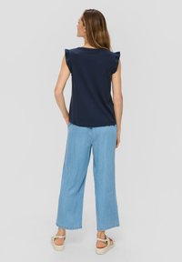 Granatowa bluzka z krótkim rękawem i falbaniastymi ramionami, zestawiona z jasnoniebieskimi szerokimi spodniami dżinsowymi oraz beżowymi sandałami na koturnie.