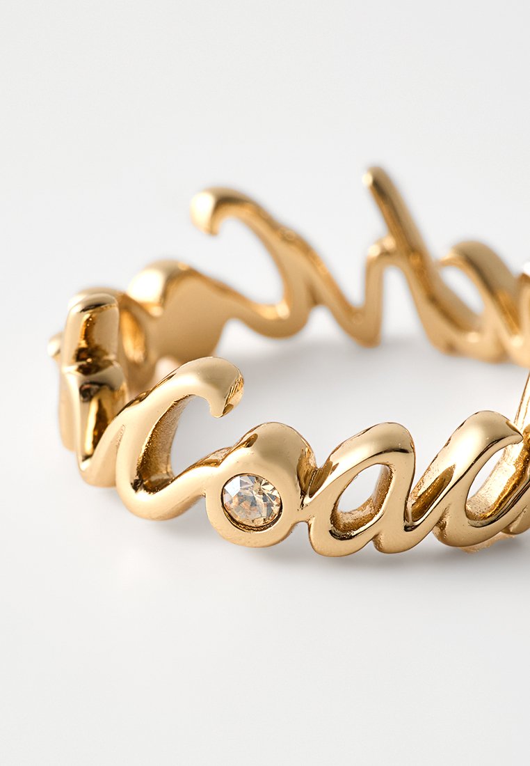 Goldring in Schreibschriftform mit einem klaren Edelstein, der in einem der Buchstaben gesetzt ist, auf weißem Hintergrund.