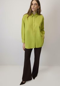 Camicia button-up oversize verde lime con colletto, abbinata a pantaloni neri svasati. La camicia presenta una texture liscia e una vestibilità rilassata.