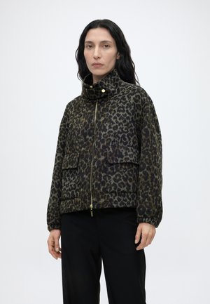 Femme aux longs cheveux noirs portant une veste zippée à col haut imprimé léopard et un pantalon noir, debout devant un fond clair uni.