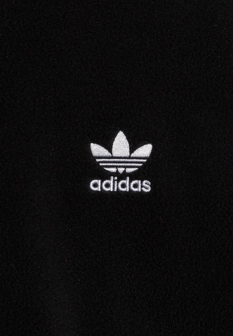 Tissu en polaire noir avec un logo Adidas brodé en blanc, composé d'un design à trois feuilles avec trois bandes horizontales en dessous.