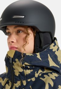 Schwarzer Helm mit grauen Akzenten, getragen mit einer Camouflagejacke, die marineblau und khakifarbene Muster aufweist. Nahaufnahme des Gesichts, die strukturierte Materialien zeigt.