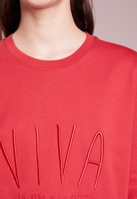 Zblízka osoba oblečená v červeném tričku s vyšitými velkými písmeny "NIVA" a menším textem pod ním na jednobarevném pozadí.