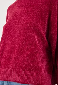 Pull en tricot rouge avec une surface texturée et un ourlet côtelé. La vue en gros plan met en valeur le matériau doux et le motif subtil.