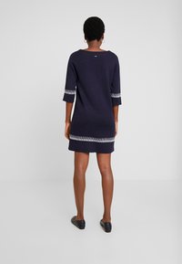Una donna con capelli corti e ricci indossa un vestito blu navy con dettagli a righe sulle maniche e sull'orlo, in piedi contro uno sfondo bianco semplice.