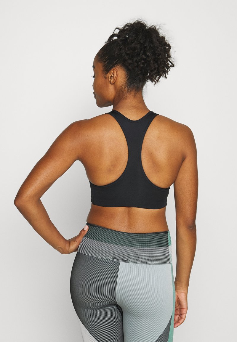 Soutien-gorge de sport noir avec design dos nageur, fabriqué en tissu extensible. Associe à des leggings à motifs dans différentes nuances de vert et de gris.