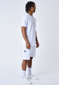 Camisa blanca de manga corta con panel frontal contrastante, combinada con pantalones cortos blancos hasta la rodilla que tienen bolsillos laterales. Zapatillas blancas y calcetines.