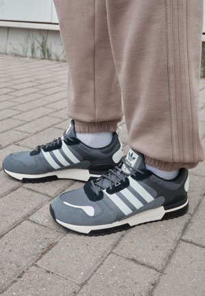 Chaussures de sport grises avec un design blanc à trois bandes, dessus en mesh texturé, col rembourré et semelle en caoutchouc noire, associées à un pantalon de survêtement beige.