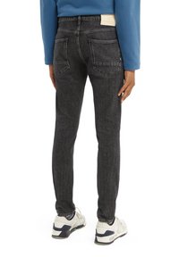 Jeans de mezclilla negra con corte slim, que presentan bolsillos traseros y un acabado texturizado. Llevados con zapatillas blancas sobre un fondo liso.