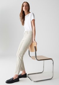 Lichtbeige corduroy broek en een aansluitend wit T-shirt, gecombineerd met zwarte instappers, terwijl hij een houten stoel met een metalen frame vasthoudt.