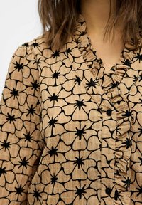 Blusa beige con un patrón floral bordado en negro, con un escote con volantes y cierre de botones, hecha de una tela texturizada.