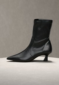Botte en cuir noir mi-mollet avec bout pointu et petit talon kitten bas, exposée sur une surface beige clair sur fond gris uni.