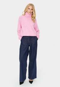 Rožinė golfas, pagamintas iš minkštos mezginio medžiagos, derinamas su laisvai krentančiomis tamsių džinsų plačiomis kelnėmis, turinčiomis dvi priekinės kišenes.