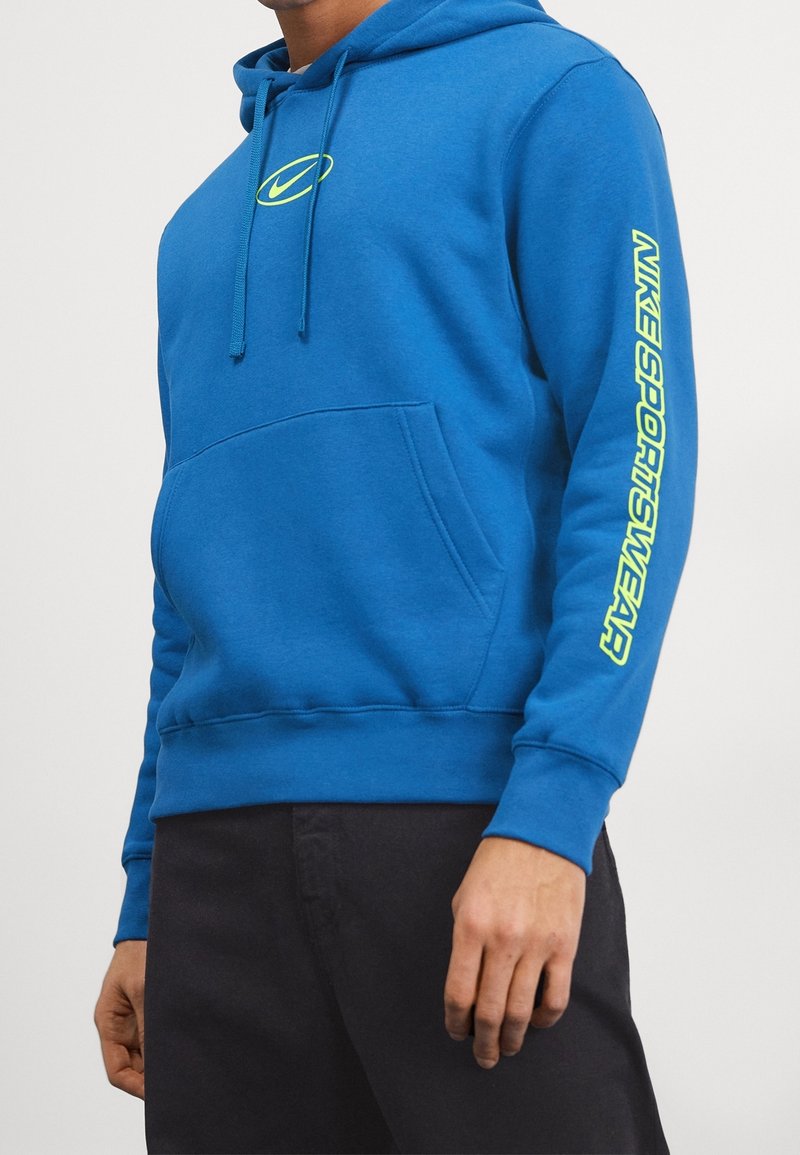 Modrá mikina s kapucí vyrobená z měkké tkaniny, s přední kapsou, šňůrkami a neonově žlutým textem Nike Sportswear podél rukávu.