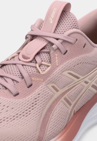 Pink athletic shoe featuring a mesh upper, contrasting beige accents, flat laces, a cushioned sole, and a loop tab for easy wear.