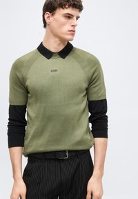 Νεαρός άνδρας που φοράει πουλόβερ με κοντά μανίκια σε χακί χρώμα πάνω από μαύρο πουκάμισο με μακριά μανίκια, μαύρο παντελόνι με ρίγες και μαύρη ζώνη.