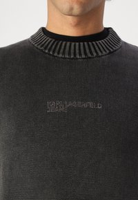 Svart stickad tröja med ribbat krage, subtil logotypdetalj och mjuk textur, designad för en tight passform runt nacken.