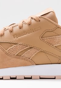 Tan Reebok-sneaker med suède-texturdetaljer, präglad logotyp, textilsnören och tjock vit sula mot en vit bakgrund.