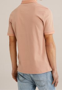 Mężczyzna w jasnoróżowej koszulce polo z krótkim rękawem w wzory oraz jasnoniebieskich dżinsach, widziany od tyłu na neutralnym tle.