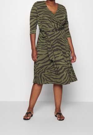 Kvinde iført knælængde olivengrøn og sort zebra-print slå-om kjole med trekvart ærmer og sorte flade sandaler.