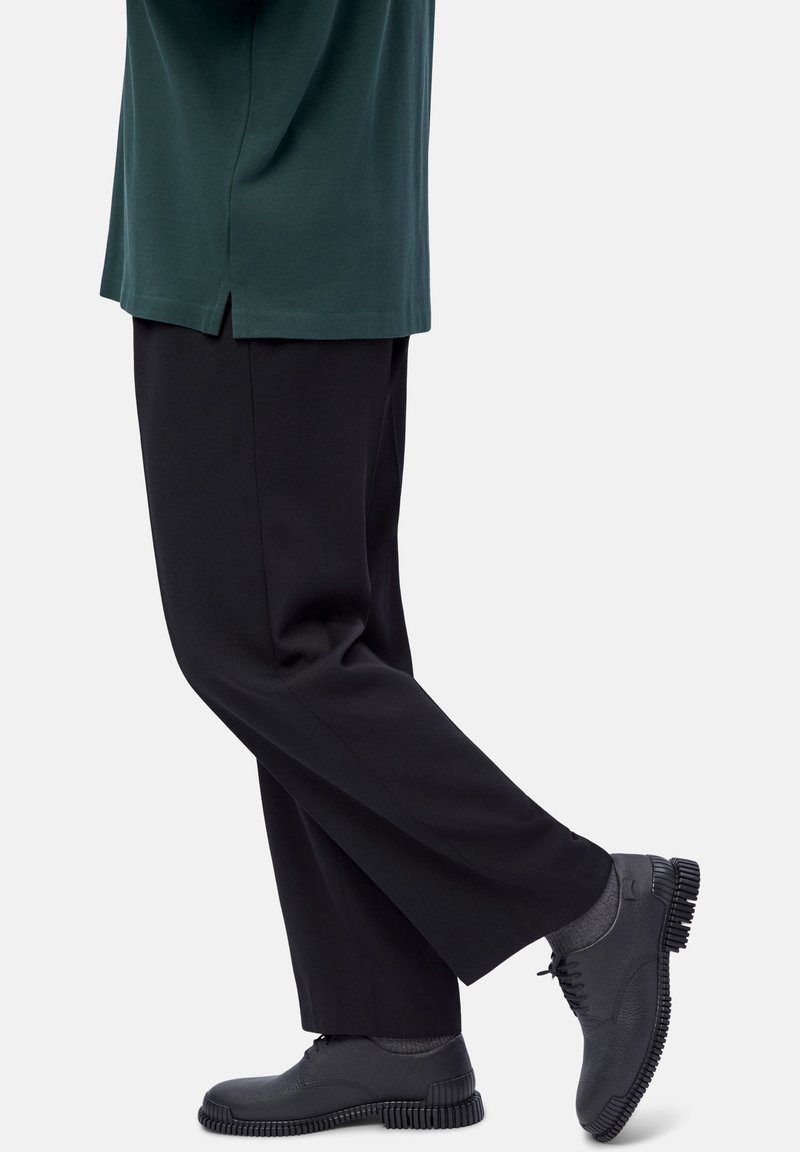 Persona che indossa pantaloni neri, calze grigie scure, scarpe stringate nere con texture e una camicia verde scuro, in piedi con un piede leggermente sollevato.