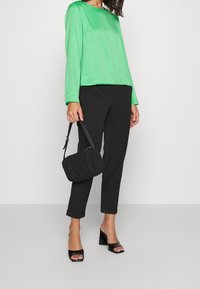 Top lungo a maniche lunghe in satin verde abbinato a pantaloni neri a sigaretta, scarpe nere lucide con tacco blocco, e una piccola borsa nera dalla texture.