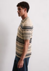 Beżowa dziana koszulka polo w poziome granatowe paski, z krótkimi rękawami i fakturowanym wzorem. Posiada kołnierzyk.