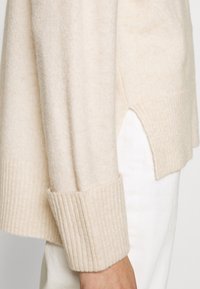 Beige stickad tröja med ribbade ärmslut och sidospel, som visar en mjuk textur och en avslappnad passform. Tyget verkar varmt och mysigt.