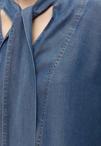 Haut en denim bleu avec des coutures surpiquées et un détail noué au niveau du cou, laissant apparaître une partie de la poitrine et du cou.