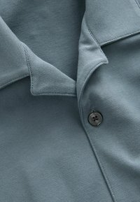 Baumwoll-Polohemd in einer gedämpften blauen Farbe mit einer strukturierten Oberfläche. Verfügt über einen spitzen Kragen und einen einzelnen dunklen Knopf an der Knopfleiste.