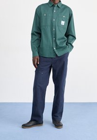 Grön knappar-upp skjorta med två bröstfickor, ihop med mörk marinblå vida byxor. Modellen bär svarta sneakers på ett ljusblått golv.