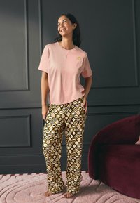 Šviesiai rožinė trumpomis rankovėmis marškinėliai su mažyte auksine lūpų grafika, derinami su raštuotomis kelnėmis, kuriose yra geometrinių formų rudos ir kreminės spalvos.
