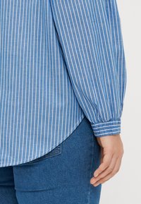 Camisa de mangas compridas azul e branca aos riscas, com a bainha arredondada e punhos franzidos, combinada com jeans azuis. Tecido texturizado visível.