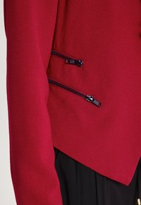 Blazer rosso con una finitura testurizzata, dotato di due tasche con zip angolate sulla parte anteriore e orlo appuntito. Le zip sono nere e lisce.