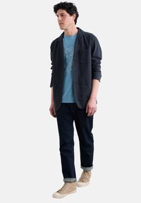 Blazer navy, camiseta de color azul claro con estampado de peces, jeans oscuros y zapatillas altas beige. Diseño en capas con ajuste casual y puños enrollados.
