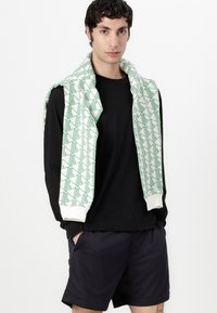 Chemise noire à manches longues associée à un short sombre. Pull vert et blanc à motifs drapé sur les épaules, avec des designs géométriques et une texture douce.