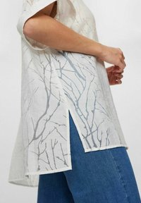 Hvid tunika med en transparent tekstur, der har et lyst grågrenmønster, korte ærmer og sideslids. Sammen med blå denim.