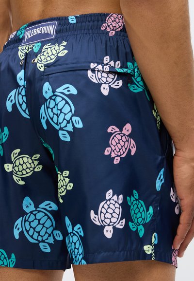 Ναυτικές σορτς κολύμβησης με πολύχρωμα σχέδια θαλάσσιων χελωνών, τσέπη με φερμουάρ στο πίσω μέρος, ελαστική μέση, φορεμένα από ένα άτομο που στέκεται με το χέρι χαλαρό στο πλάι.