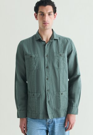 Hombre con cabello corto y rizado que lleva una camisa verde abotonada con cuatro bolsillos delanteros y vaqueros azul claro, de pie frente a un fondo liso.