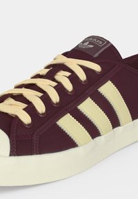 Μπορντό αθλητικά παπούτσια Adidas με κρεμ κορδόνια και ρίγες, υφασμένη λευκή καουτσουκέ πέλμα και λογότυπο στη γλώσσα.