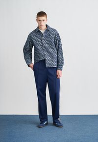 Modra srajca s potiskom in z zapenjanjem na gumbe, kombinirana s temno modrimi športnimi hlačami z belimi črtami. Model stoji na modri teksturirani podlagi.