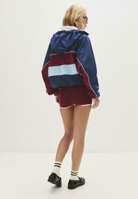 Veste coupe-vent colorée avec un design bleu marine, bordeaux et bleu clair, dotée d'une capuche et de poignets élastiques, assortie à un short bordeaux et des chaussures noires.