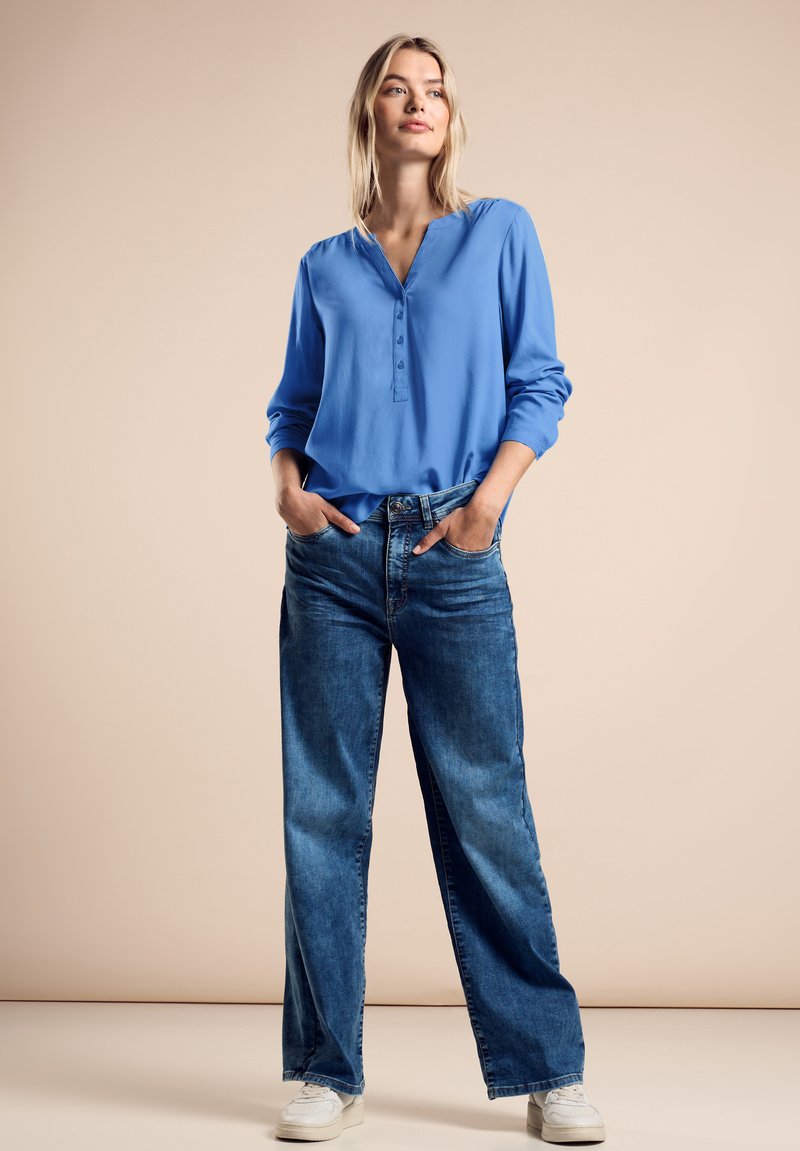 Ljusblå blus med lång ärm och v-ringning, med knapplist, kombinerad med löst sittande blå denimjeans och vita sneakers.