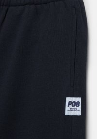 Ciemnogranatowe spodnie dresowe z elastycznym pasem i boczną kieszenią, z białą metką z napisem "P08 Studio Rotterdam".