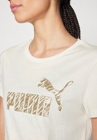 Kvinna iklädd en vit t-shirt med en beige zebrarandig Puma-logotyp över bröstet, med flätat hår uppsatt.