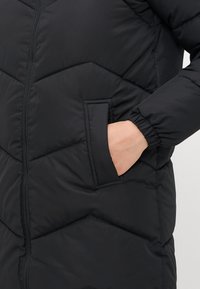 La mano di una persona inserita nella tasca laterale di una giacca nera trapuntata con cerniera e polsino elasticizzato sulla manica.